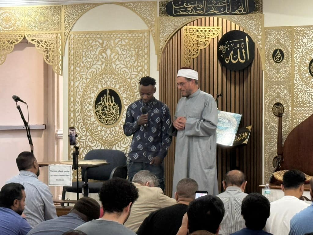 A young man speaking into a microphone beside the Imam at the front of the prayer hall, the congregation seated before them